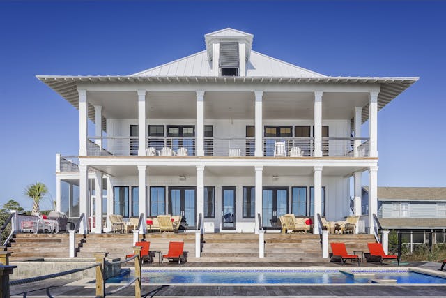 A modern, white two-story luxury home with a pool and multiple lounging areas under clear blue skies