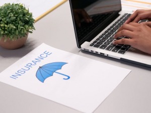 A person typing on a laptop, a piece of paper with a blurred umbrella, and the word "Insurance" on the table.